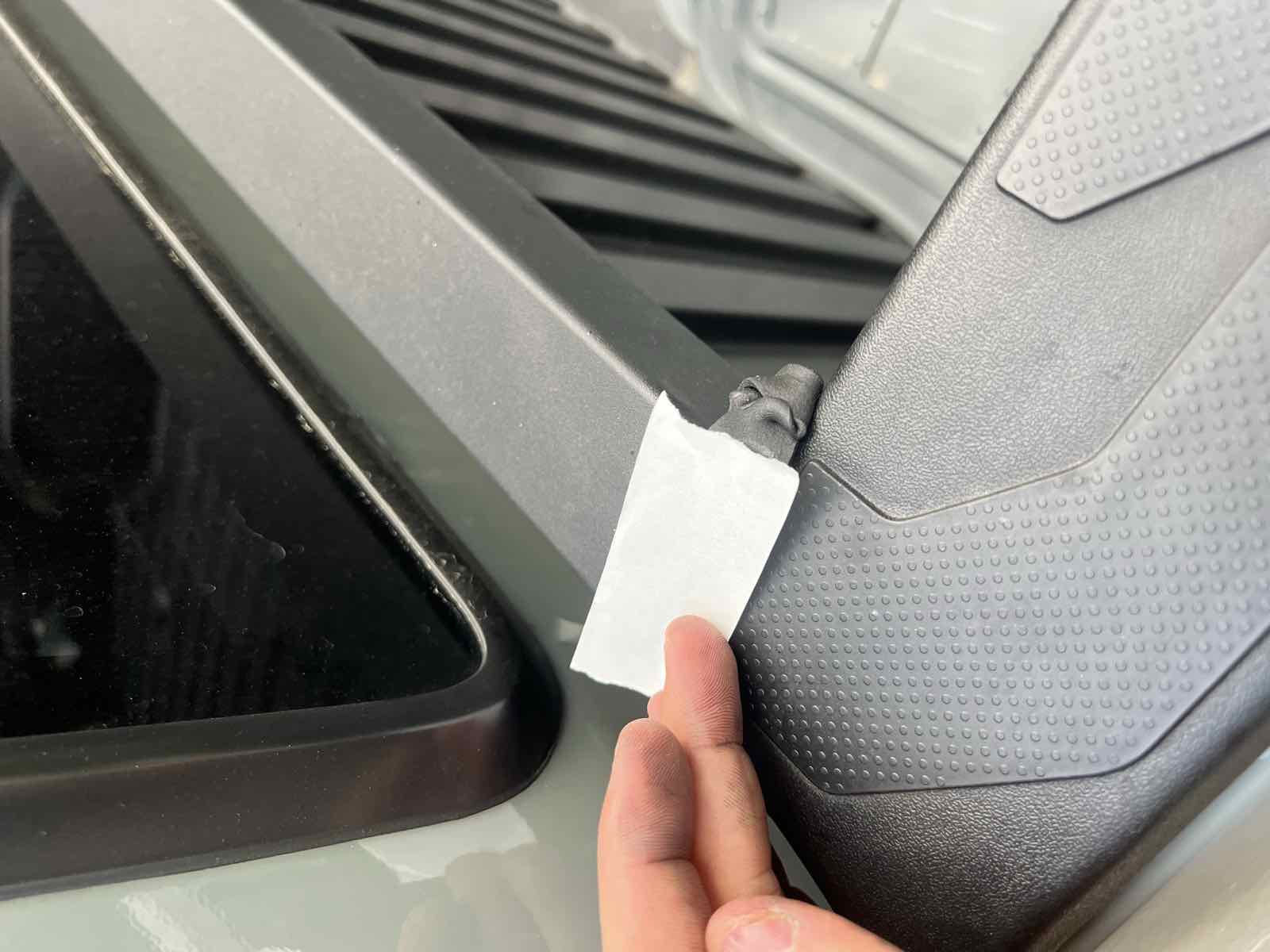 A hand pressing butyl seal tape into the front bed-rail corner of a Tacoma using the wax-paper backing strip so the sticky butyl does not stick to fingers.