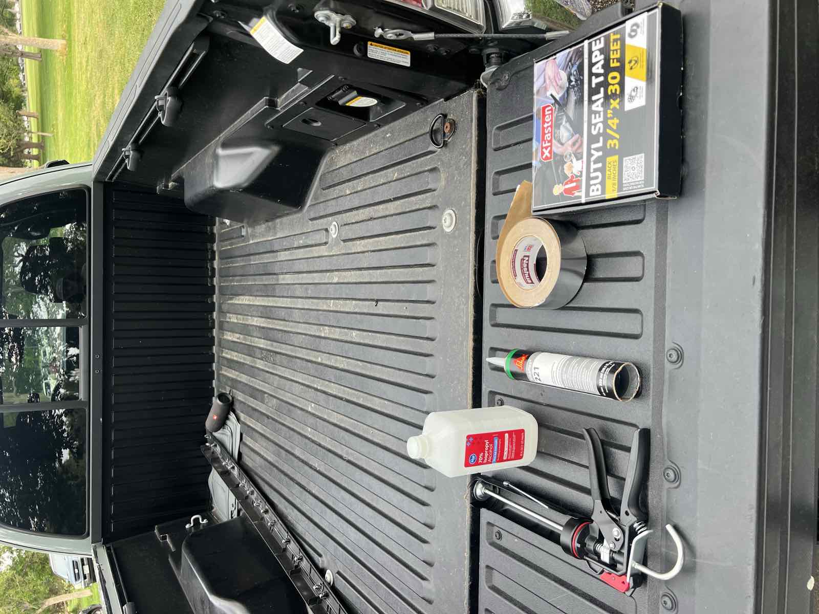 Bed-sealing supplies laid out on a 2023 Tacoma tailgate: a caulk gun, 70% isopropyl alcohol, Sikaflex 221, a roll of Nashua aluminum foil tape, and XFasten butyl seal tape, with the empty truck bed behind.