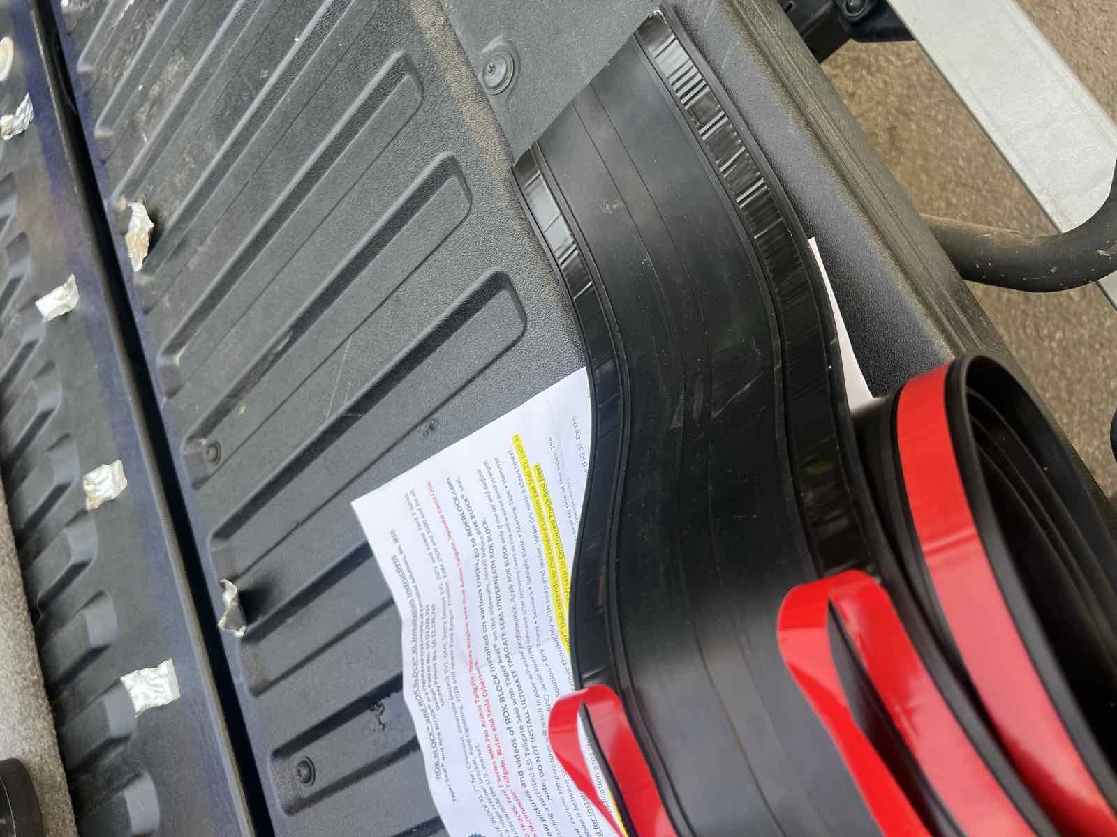 A hand peeling back a short section of red adhesive liner from a ROK Block seal partway along a Tacoma tailgate, keeping the remaining liner attached.